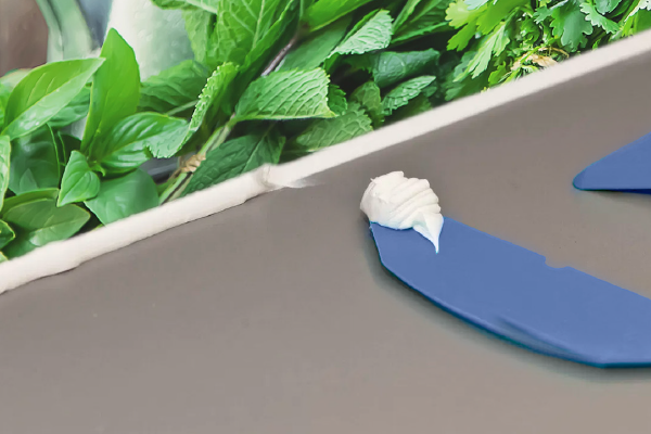 Nahaufnahme eines blauen Spachtels, der Dichtmasse auf Tisch aufträgt, mit frischen Kräutern im Hintergrund